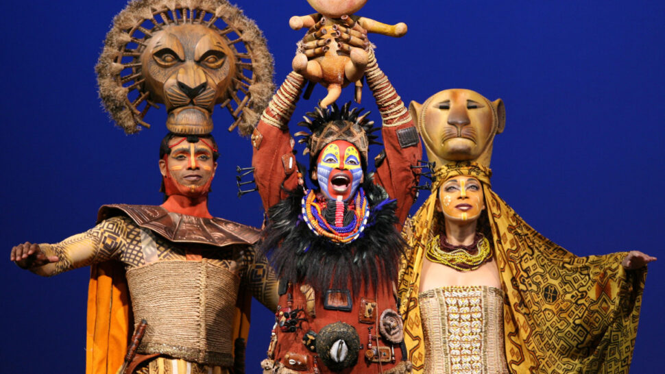 A frontal shot of three characters in Lion King wearing colorful costumes. The character to the left has an orange cape and wears a symbol of the sun on his head. The figure in the middle has orange, yellow and blue stripes on their face while holding up in both hands above their head a lion cub. The character to the right wears a lion mask on top of her head (with her face showing). From the left to right: Mufasa (played by Nathaniel Stampley), Rafiki (by Tshidi Manye), and Sarabi (by Jean Michelle Grier) in Liong King. Costume design by Julie Taymor.